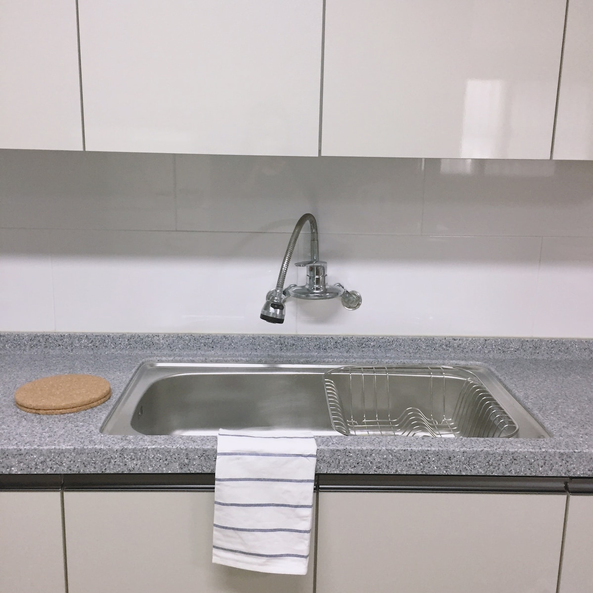 A clean kitchen sink area features a stainless-steel sink with a modern faucet. A drying rack is placed beside the sink, with a dish towel draping over the edge. The countertop is finished in a polished gray stone, complementing the white cabinetry above.