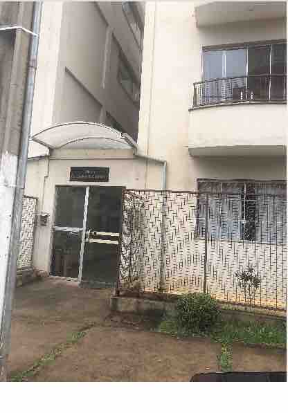 A building entrance is depicted, featuring a glass door under a metal canopy. The exterior showcases a simple design with concrete walls and nearby windowed balconies. A low fence and some greenery are present in the surrounding area, indicating a well-maintained environment.