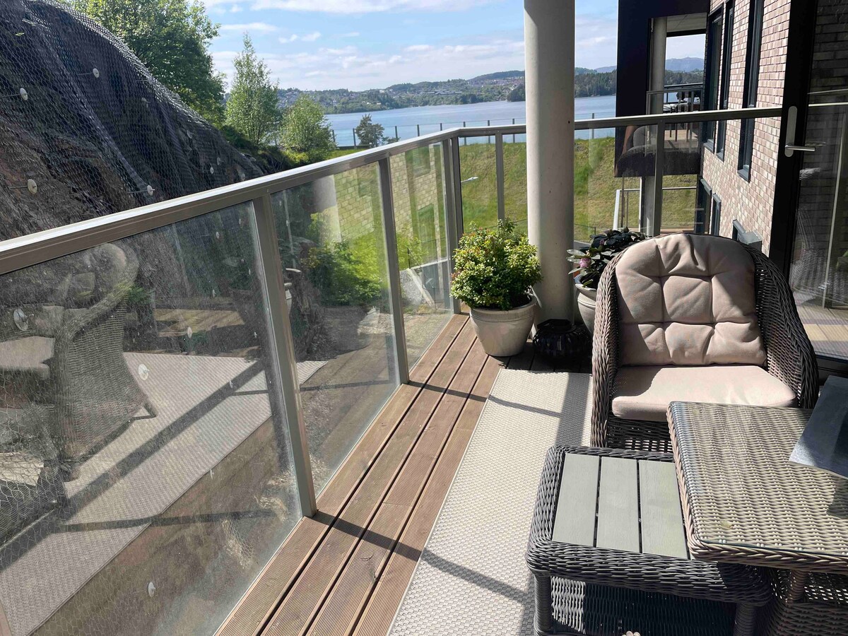 A glass-walled terrace offers views of the water and surrounding greenery. Outdoor seating is provided with a comfortable chair and a small table. Potted plants add a touch of greenery to the space, enhancing the serene atmosphere.