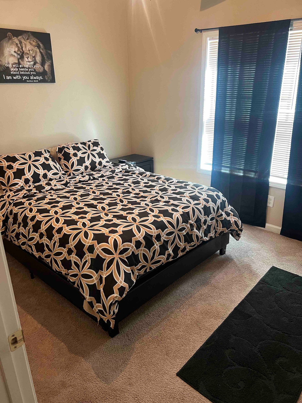 A cozy sleeping area features a black and white floral patterned duvet covering a spacious bed. Natural light enters through partially open blinds, illuminating the soft carpet. A dark rug lies beneath the bed, and a nightstand stands beside it, adding functionality to the space.