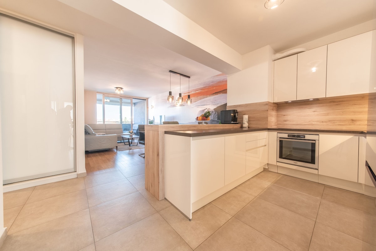 A modern kitchen space features white cabinetry and a wood-accented countertop. Equipped appliances include an oven and stovetop. Natural light floods the area from the adjacent living space, visible through large windows. The flooring is finished in neutral tiles, creating a cohesive look.