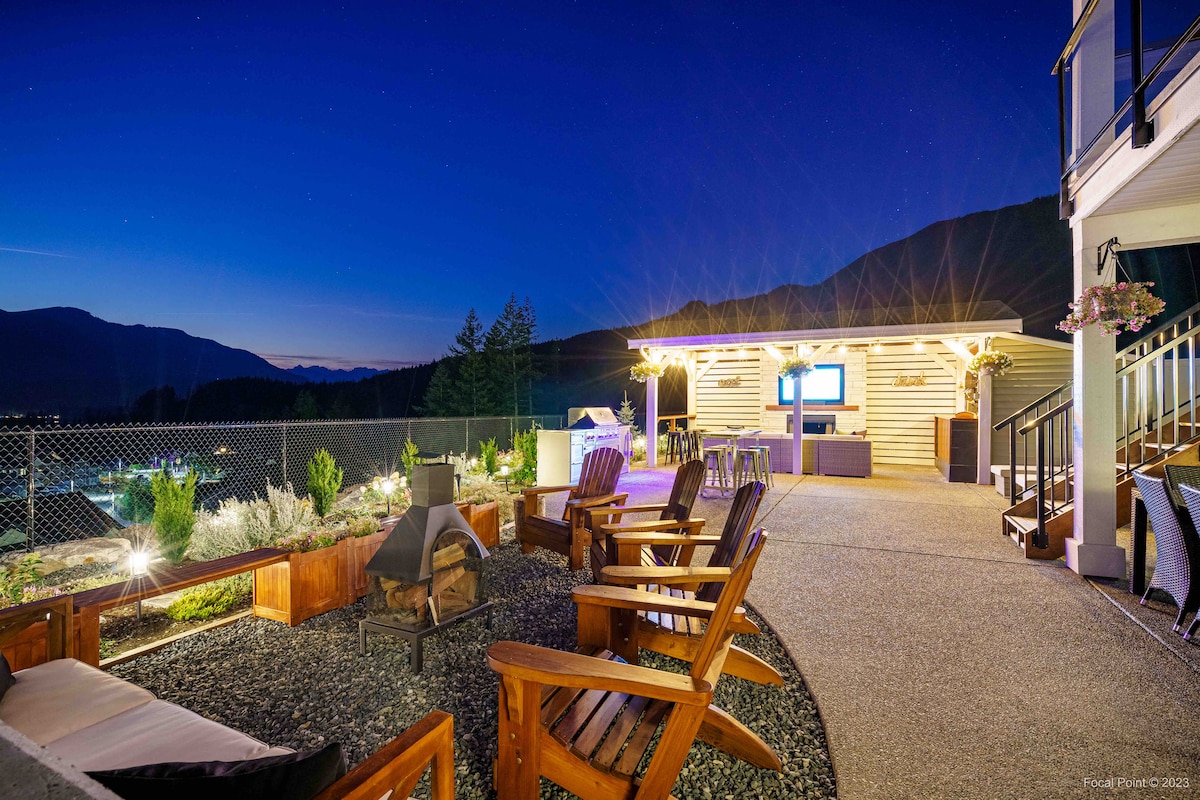 An outdoor seating area features several wooden chairs arranged around a fire pit, with a pathway leading to a beautifully lit gazebo. The space is surrounded by greenery and offers expansive views of the night sky and nearby mountains.