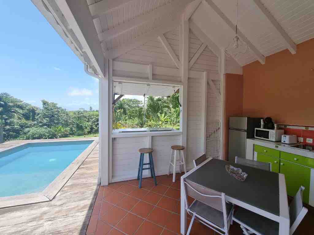 An open kitchen area features sleek cabinetry and modern appliances, including a microwave. A dining table with white chairs is positioned near large windows that provide views of the lush surroundings and the swimming pool. Natural light brightens the space, enhancing the inviting atmosphere.