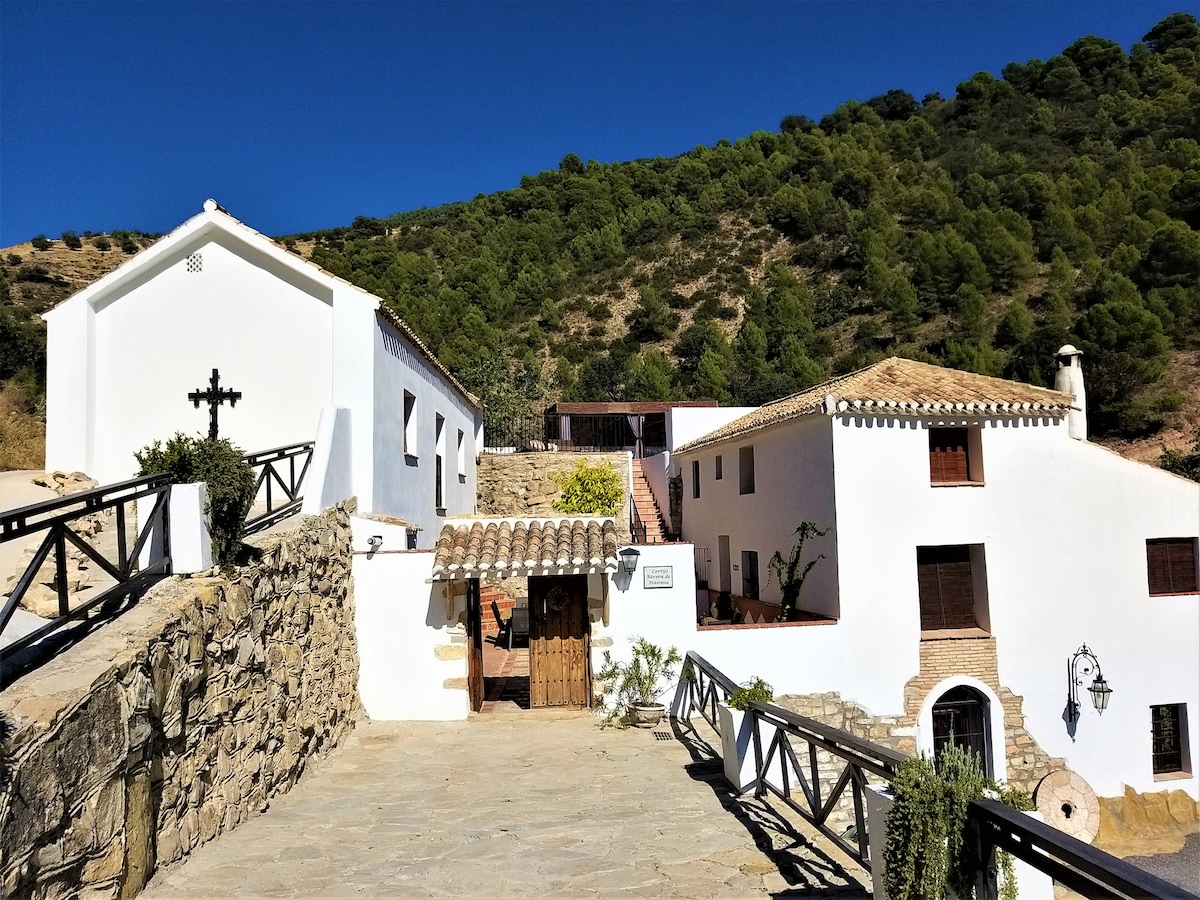 Two charming white houses are presented, set against a backdrop of lush green hills. A stone pathway leads to a wooden door, while quaint details like a cross and decorative elements enhance the welcoming entrance.
