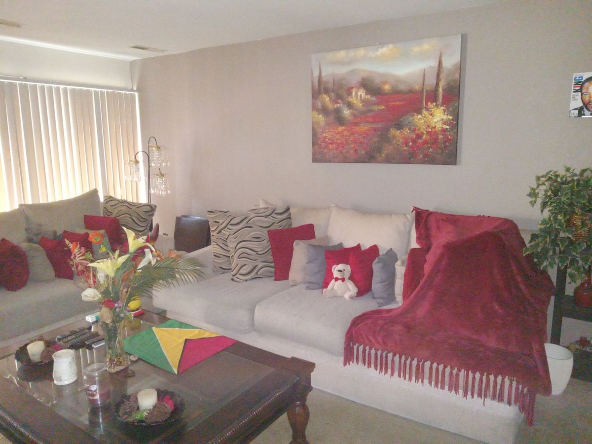 A cozy living area is presented with a spacious light-colored sectional sofa adorned with various cushions in different patterns. A plush blanket drapes over one end. A coffee table sits at the center, with candles and decorative plants adding a touch of color and comfort.