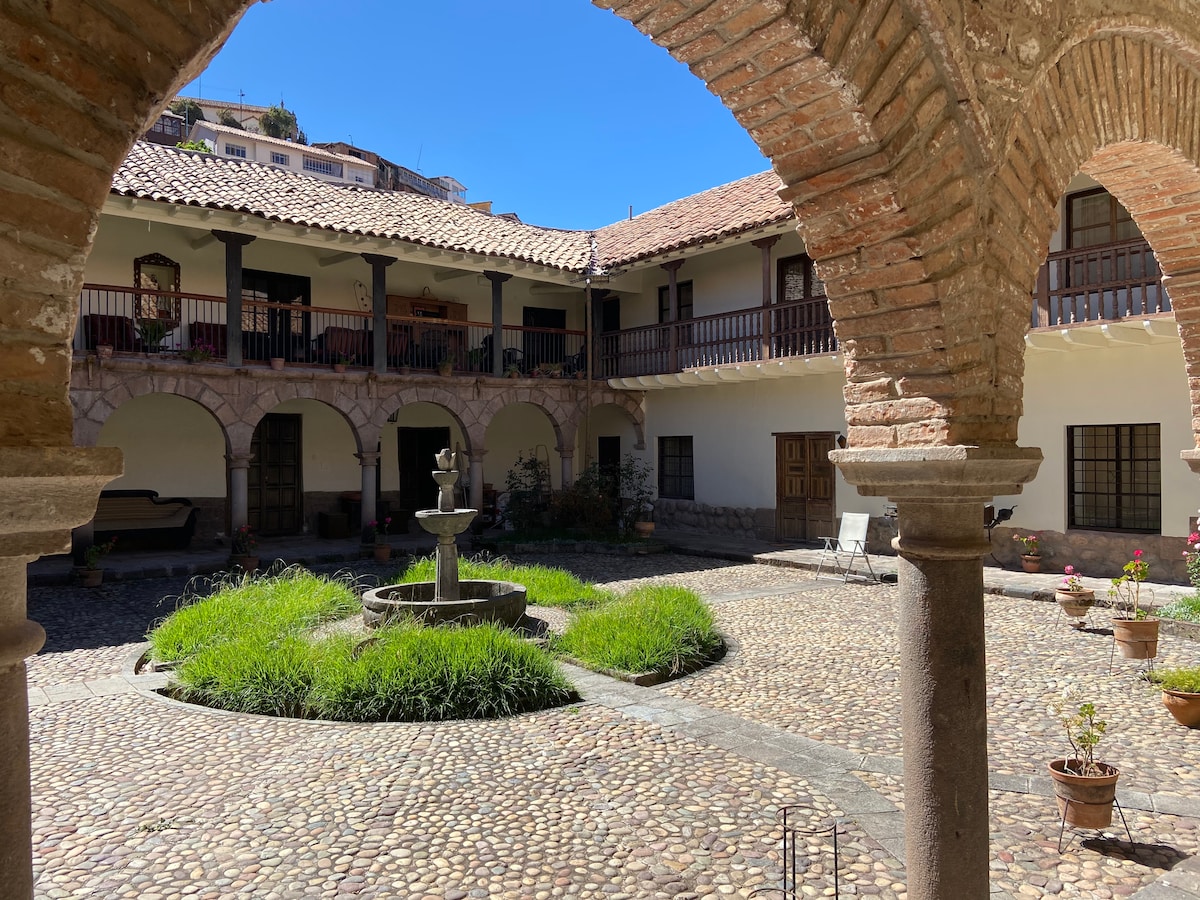 A charming courtyard is framed by arched brick columns, featuring a central fountain surrounded by lush grass. Potted plants add greenery to the cobblestone surface, and upper-level balcony spaces overlook the serene scene.