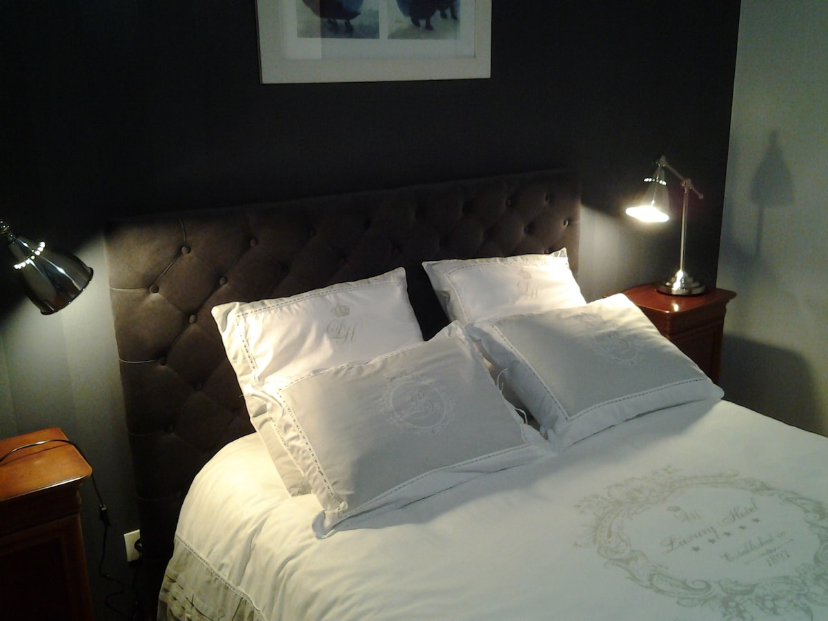 A spacious bed with an upholstered headboard is positioned along a dark wall. Soft, light-colored bedding is complemented by decorative pillows. Bedside tables are placed on either side, each featuring a lamp for additional lighting.
