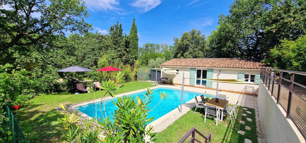 A well-kept private pool is surrounded by lush greenery, with comfortable lounge chairs positioned nearby. Tables with umbrellas provide shaded spots for relaxation. The gîte is visible in the background, framed by trees and featuring large windows for natural light.