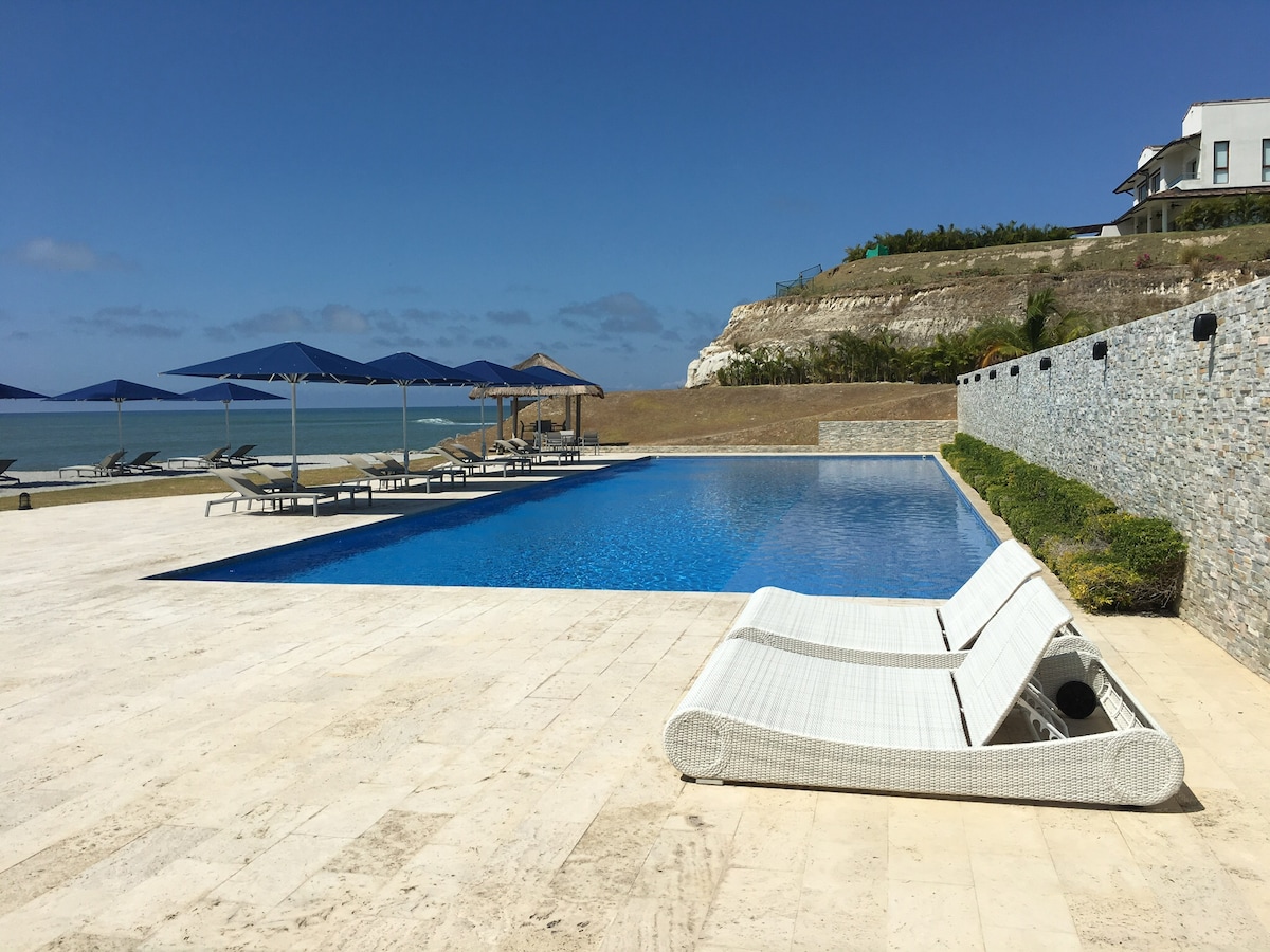 A spacious outdoor pool area is visible, featuring a large rectangular pool with clear blue water. Several lounge chairs are arranged around the pool, accompanied by sunshades providing shade. A grassy area and a stone wall frame the scene, along with a view of the ocean in the distance.