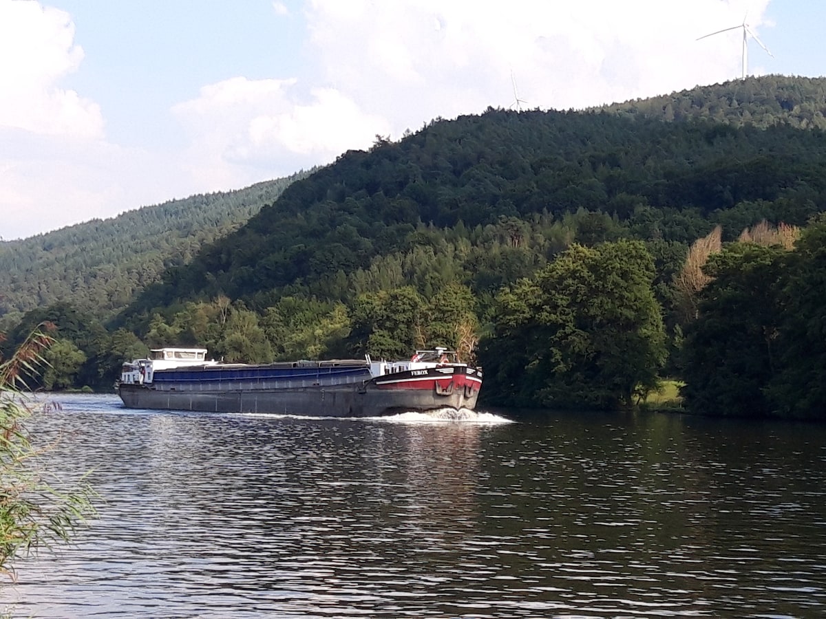 A cargo ship navigates along a calm river, surrounded by lush green hills and trees. The hills rise gently in the background, while a clear sky is dotted with fluffy white clouds. The water's surface reflects the natural scenery, creating a peaceful scene.