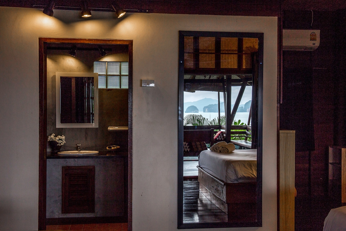 A spacious interior is captured, showcasing a modern bathroom with a sink and mirror adjacent to a bedroom area. Large glass doors frame the stunning views of Phang Nga Bay, while soft natural light illuminates the inviting space.