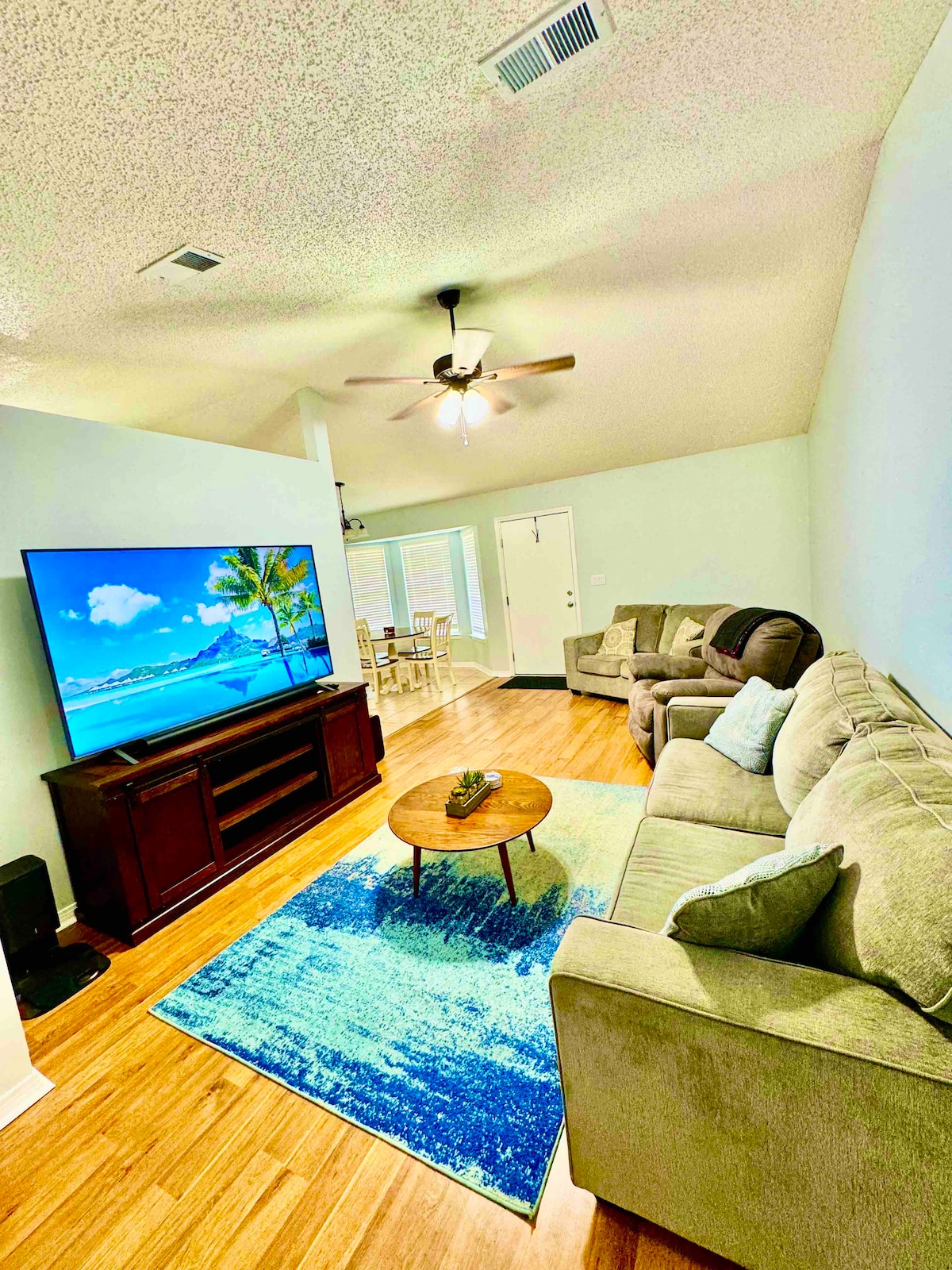 A bright living area features a large smart TV displaying a tropical scene. Comfortable seating arrangements include a light-colored sofa and two accent chairs. A round coffee table is positioned on a patterned rug, while natural light enters through nearby doors leading outside.