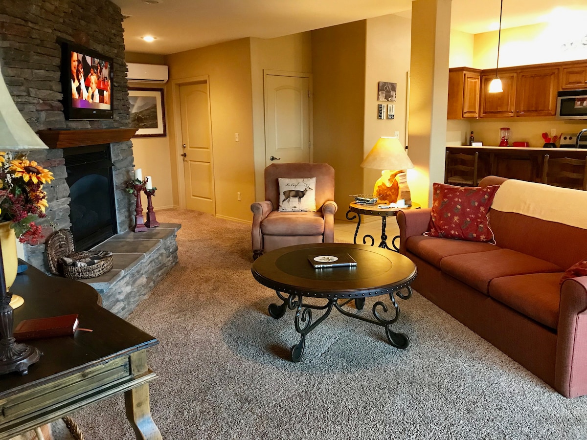 A cozy living area features a stone fireplace with a mounted television above. A large, round coffee table sits center stage, surrounded by a sofa and armchair. Soft lighting from a lamp and decorative items add warmth to the space, with a view into the kitchen area.