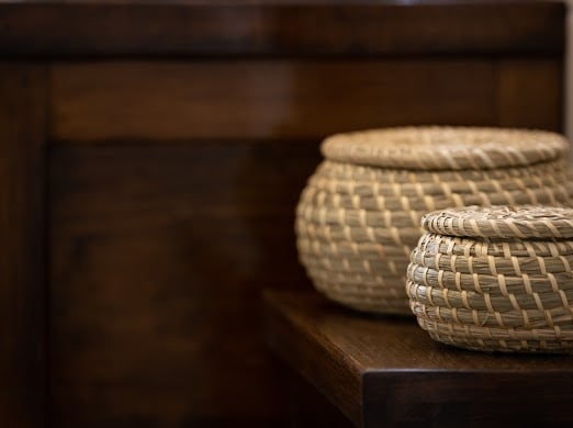 Two woven baskets of varying sizes are placed on a wooden surface. The natural texture and color of the baskets contrast with the dark wood of the surrounding furniture, highlighting a simplistic yet elegant storage option.