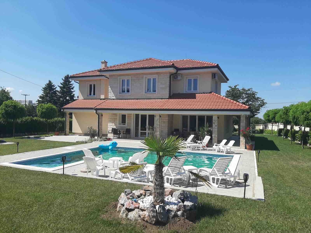 A spacious house with a tiled roof is surrounded by well-maintained grass and trees. A large pool is framed by lounge chairs, while a neatly arranged garden features a small palm tree and decorative stones.