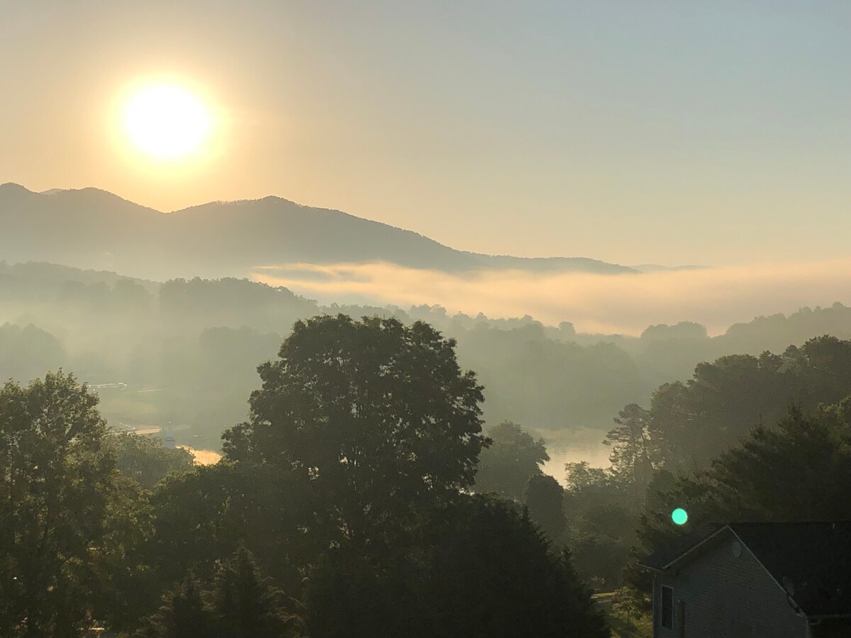 A tranquil morning scene is captured, showcasing sun rays filtering through mist across rolling hills and trees. The soft glow of sunlight illuminates the landscape, with the outlines of distant mountains visible in the background. A serene body of water reflects the surrounding nature.