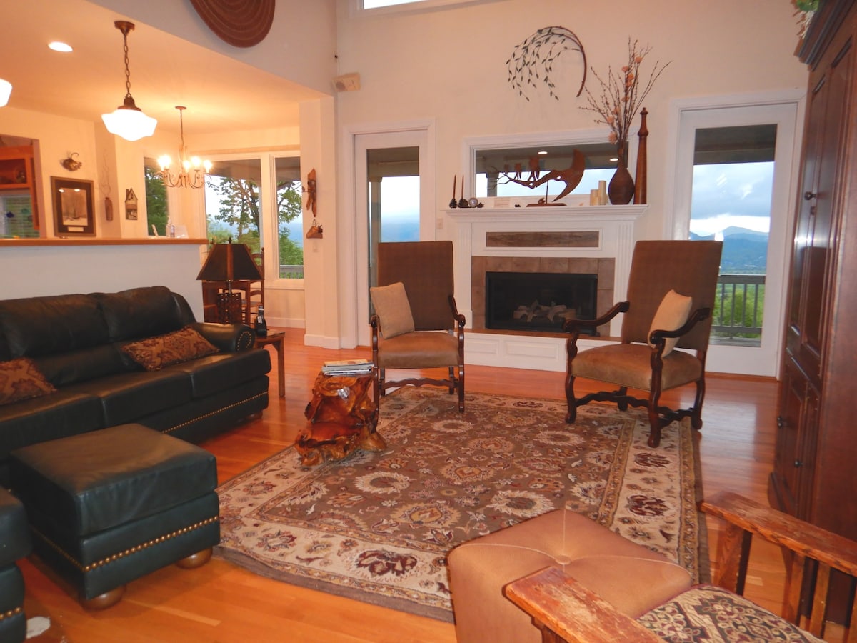 The living area is equipped with a dark leather sofa and two upholstered chairs. A decorative area rug sits on hardwood flooring, complementing the fireplace and mantel. Large windows provide scenic mountain views and natural light, enhancing the open and spacious design.