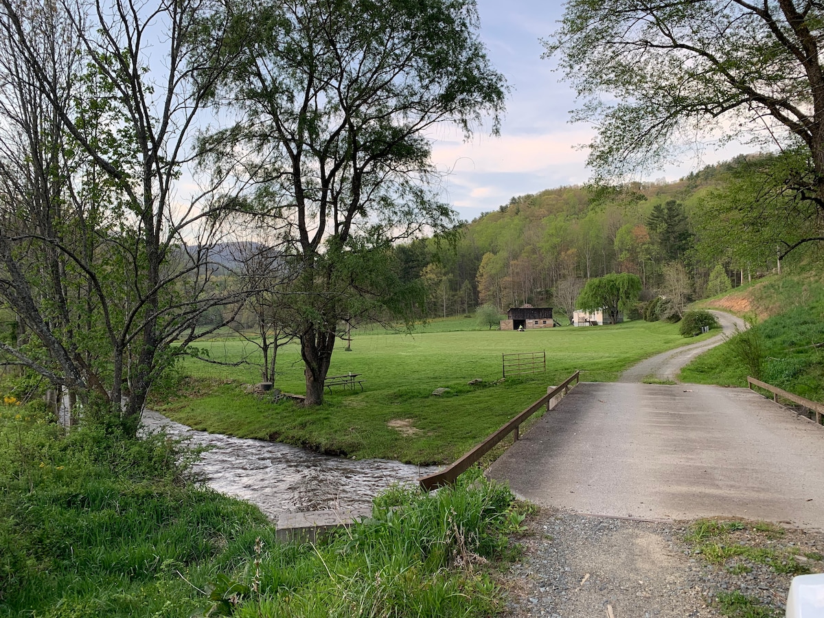 A gentle stream flows alongside a gravel road that leads to a lush green field. Trees frame the scene, with a distant view of a house nestled in the landscape and rolling hills rising in the background.