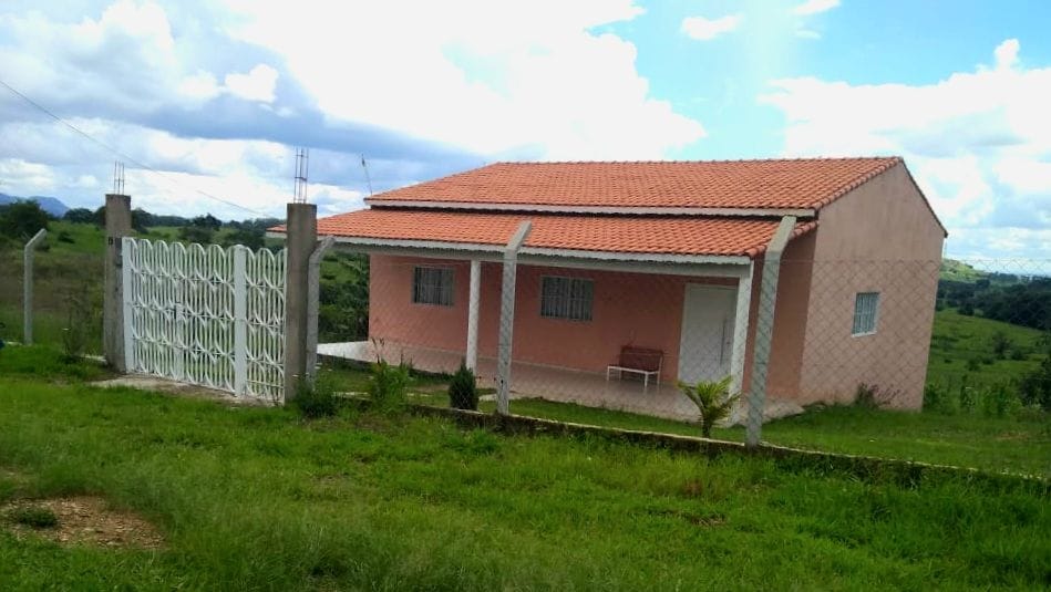A single-story house is depicted with a sloped roof and a welcoming facade. Surrounding the home, grassy areas and gentle hills are visible under a partly cloudy sky. A mesh fence outlines the property, adding a sense of security and openness to the surrounding landscape.