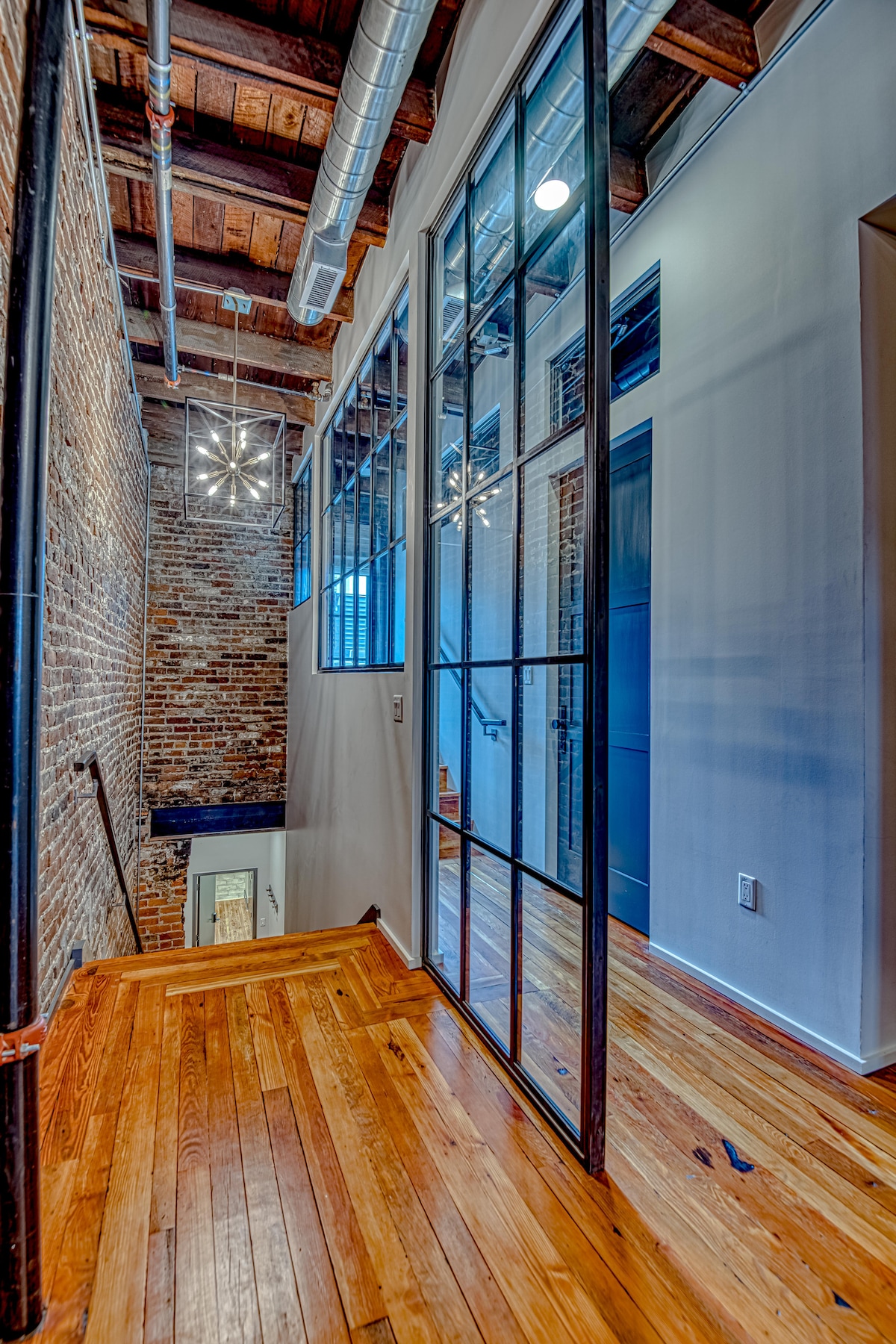 A hallway features exposed brick walls and wooden flooring. Large glass panels provide a view of the adjoining space. Suspended light fixtures enhance the contemporary design, while stairs lead down to additional areas of the condo.