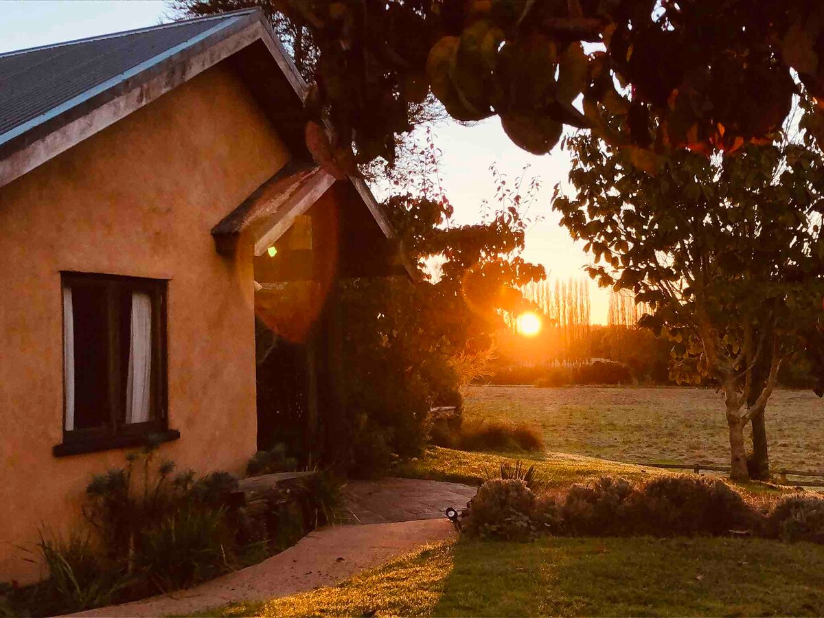 The exterior of a Mediterranean-style holiday home is viewed at sunset, with warm orange hues illuminating the adobe walls. The surrounding green landscape offers a tranquil setting, complemented by trees and a clear sky. Soft sunlight filters through the leaves, creating a peaceful atmosphere.