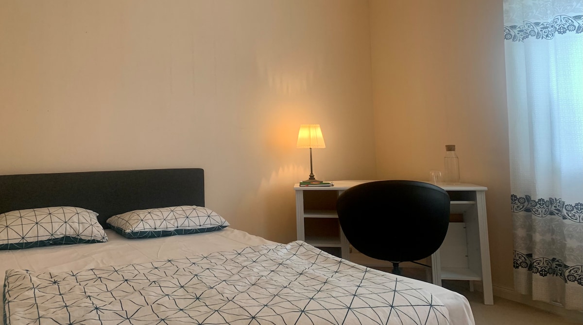 A spacious bedroom features a large bed with a simple patterned blanket and two pillows. A lamp provides soft lighting beside the bed, while a desk and chair are positioned nearby. Bright light enters through a window adorned with light curtains.