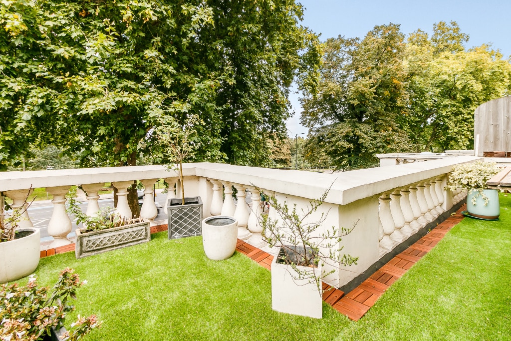 A spacious terrace is adorned with greenery and potted plants. The railing features decorative balustrades, while manicured grass creates a pleasant outdoor area. Trees can be seen in the background, providing a natural, leafy environment.