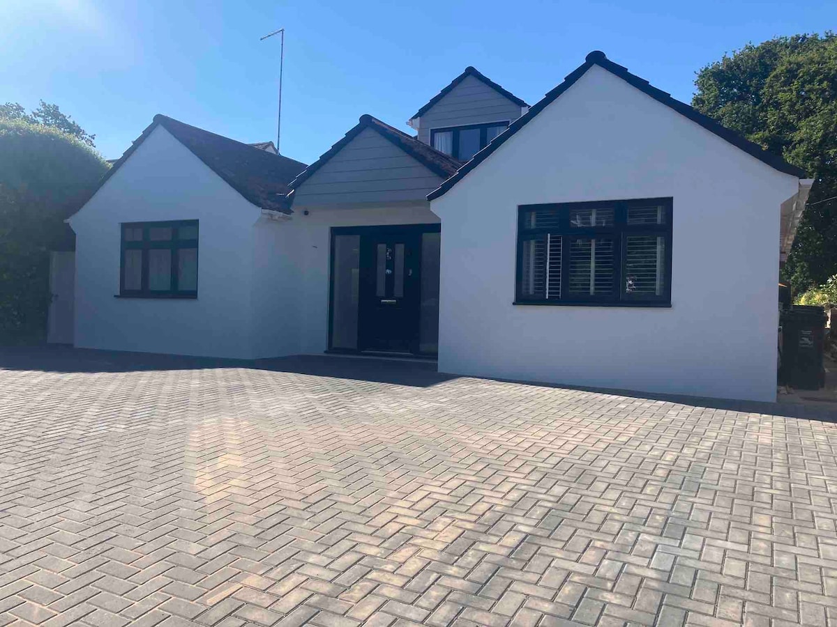 A modern two-toned house is featured, with a white exterior and dark windows. The driveway is expansive, paved with geometric patterns, ensuring ample parking space. Lush greenery borders the property, and a clear sky is overhead, enhancing the inviting appearance of the home.