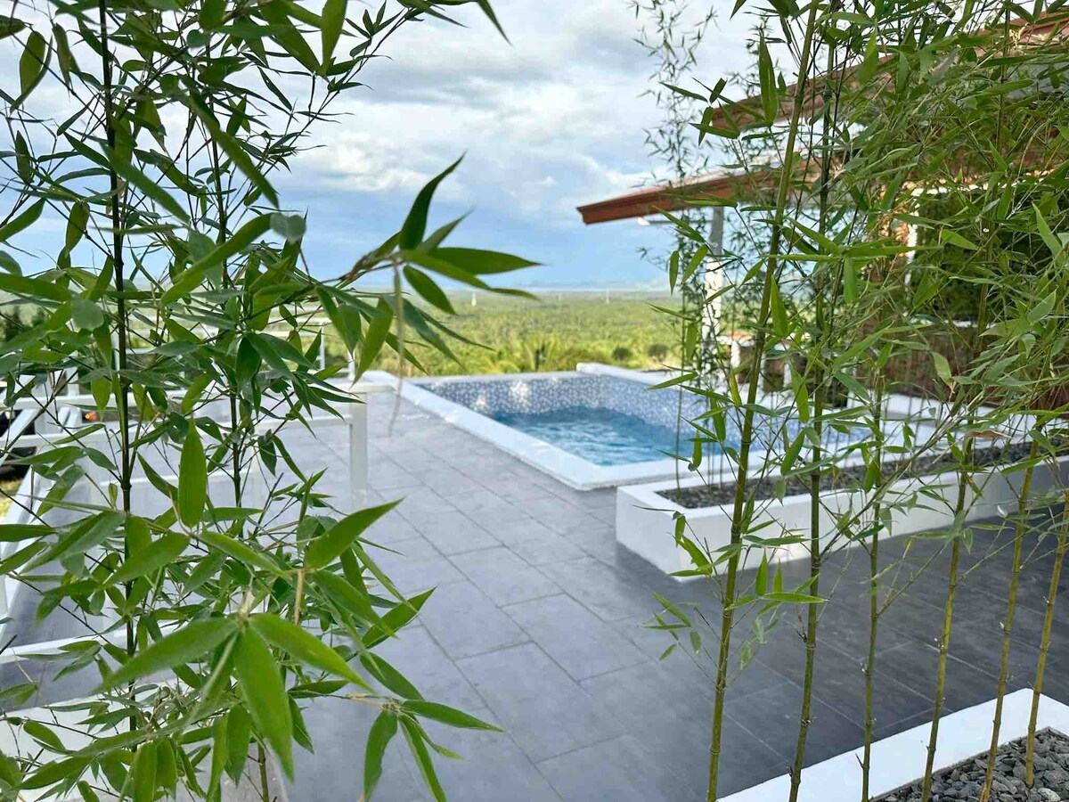 A serene outdoor area is highlighted by a swimming pool surrounded by greenery. The polished stone patio features bamboo plants framing the view, with expansive scenery visible in the background, offering a connection to nature.