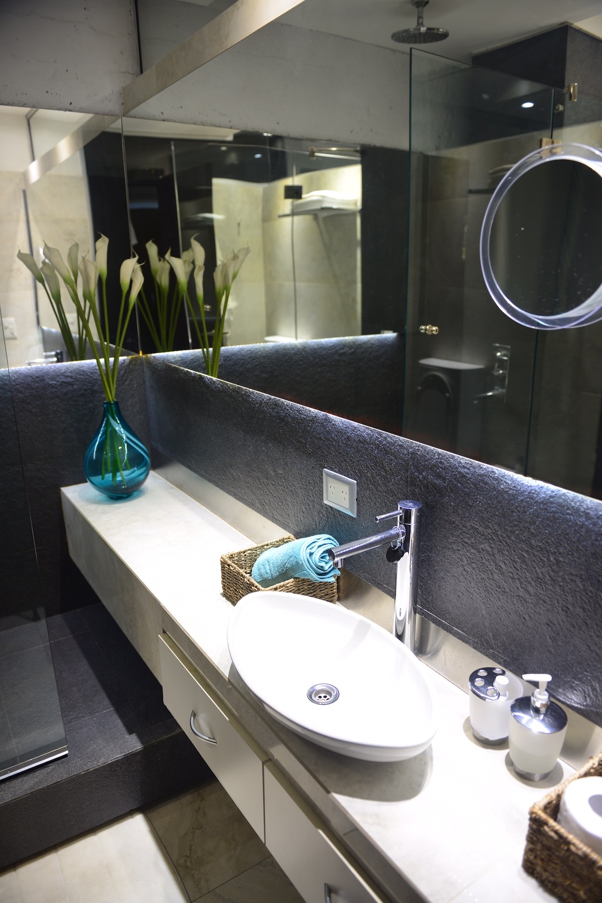 The bathroom features a sleek sink set atop a smooth countertop, complemented by a decorative blue vase filled with fresh flowers. A large mirror enhances the space, while neutral-toned tiles and modern fixtures contribute to a clean and contemporary atmosphere.