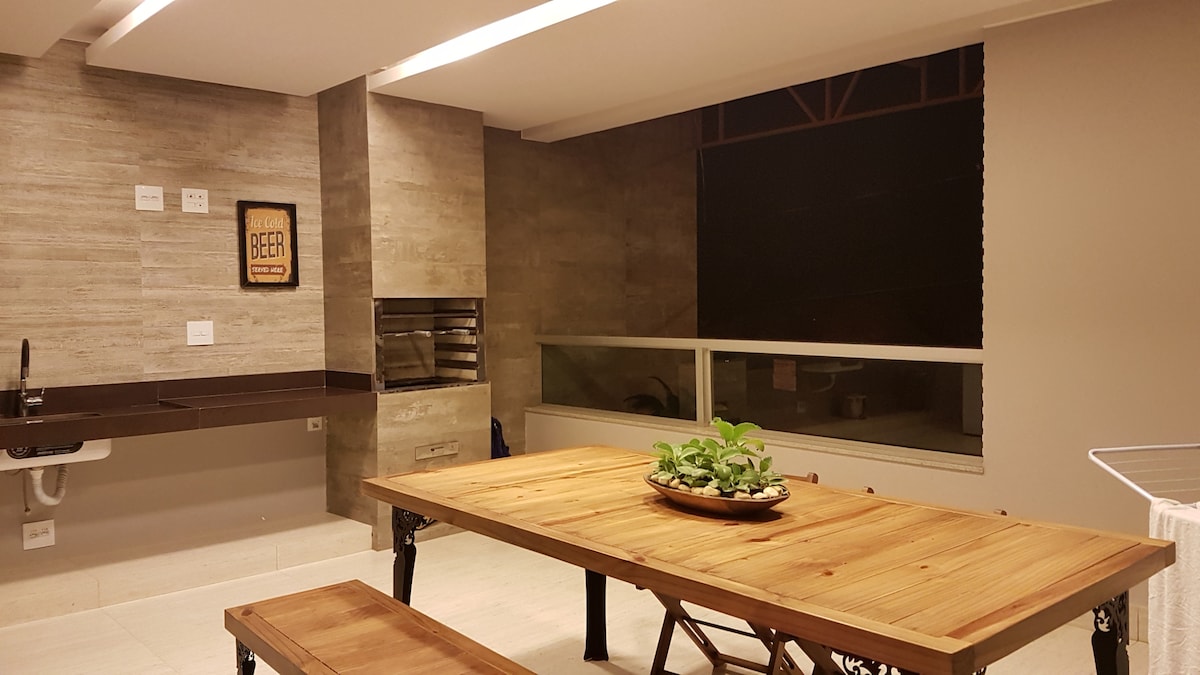 A spacious gourmet area features a large wooden table with natural finish, surrounded by bench seating. A stone wall serves as a backdrop for a grill. A large window allows for ample natural light, revealing the view outside.