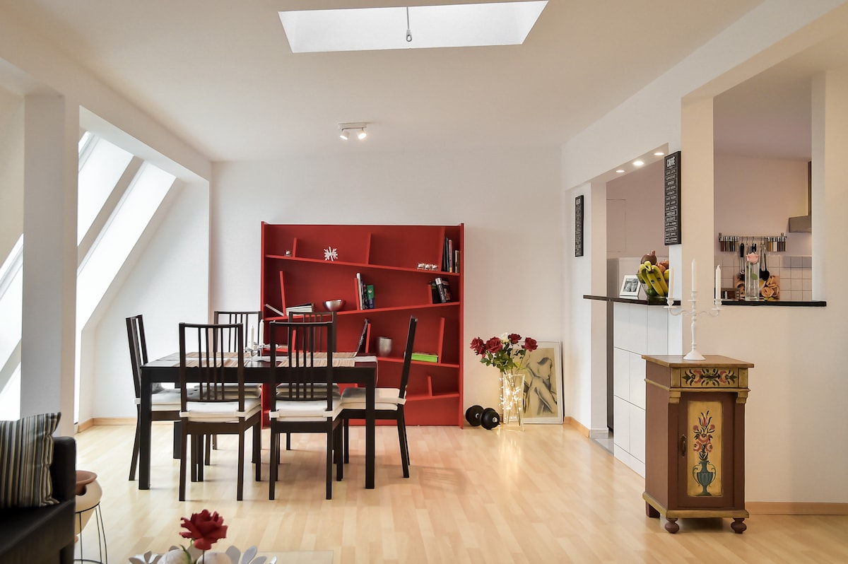 A spacious living area features an open layout with ample natural light flooding in through skylights. A modern red bookshelf adds a pop of color, complemented by a dining table surrounded by black chairs. A decorative cabinet and some plants contribute to the inviting atmosphere.