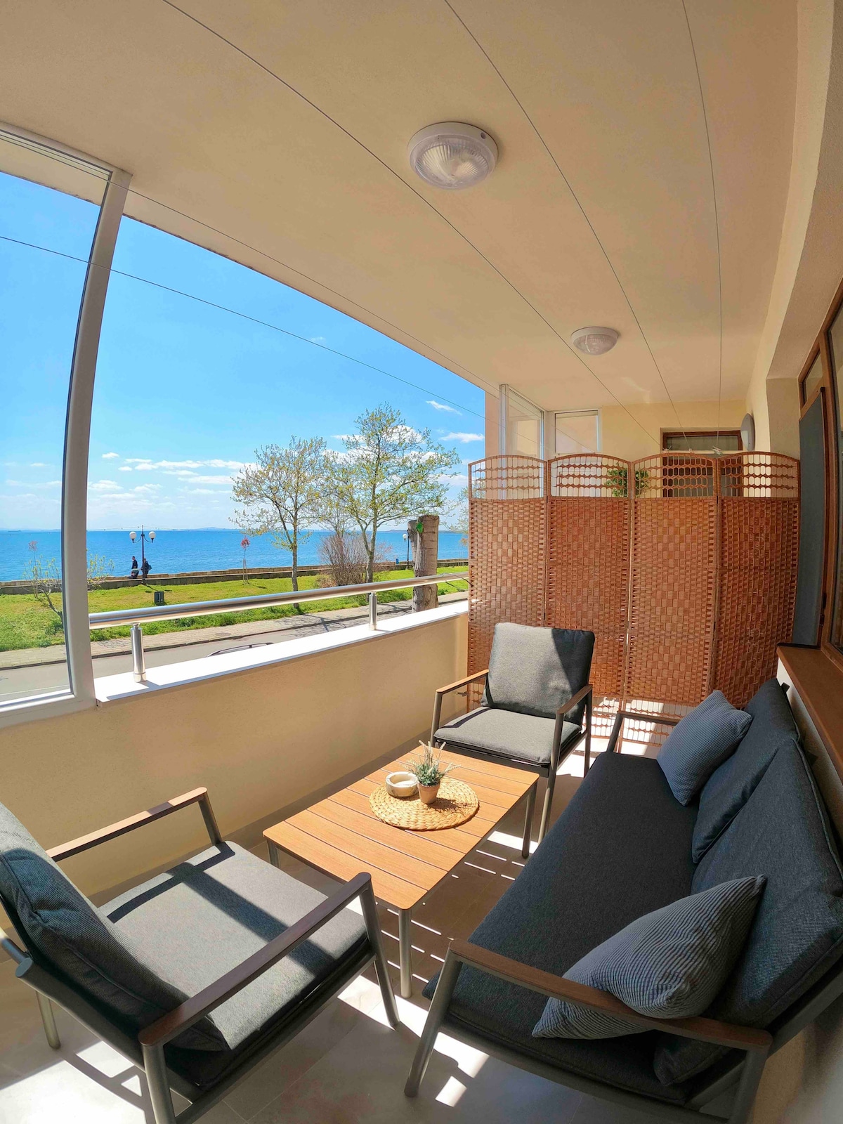 A spacious balcony offers sea views through large windows, featuring comfortable seating arrangements with grey cushions. A wooden table sits at the center, and decorative privacy screens are positioned to create a cozy atmosphere. The surrounding area includes green trees and a pathway near the water.