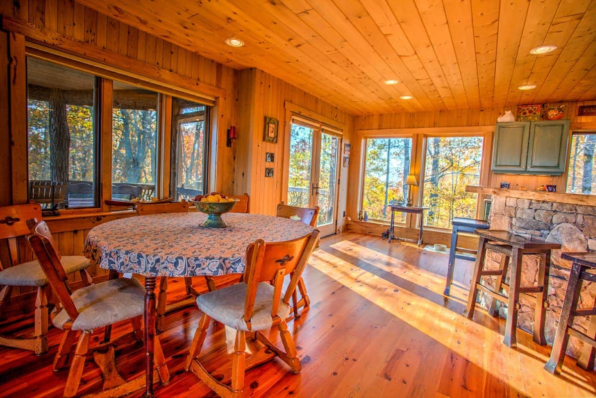 A bright dining area is highlighted by large windows that offer expansive views of the surrounding trees. A wooden table is surrounded by several chairs, and a stone fireplace is visible in the background. Warm wood paneling creates a cozy ambiance.