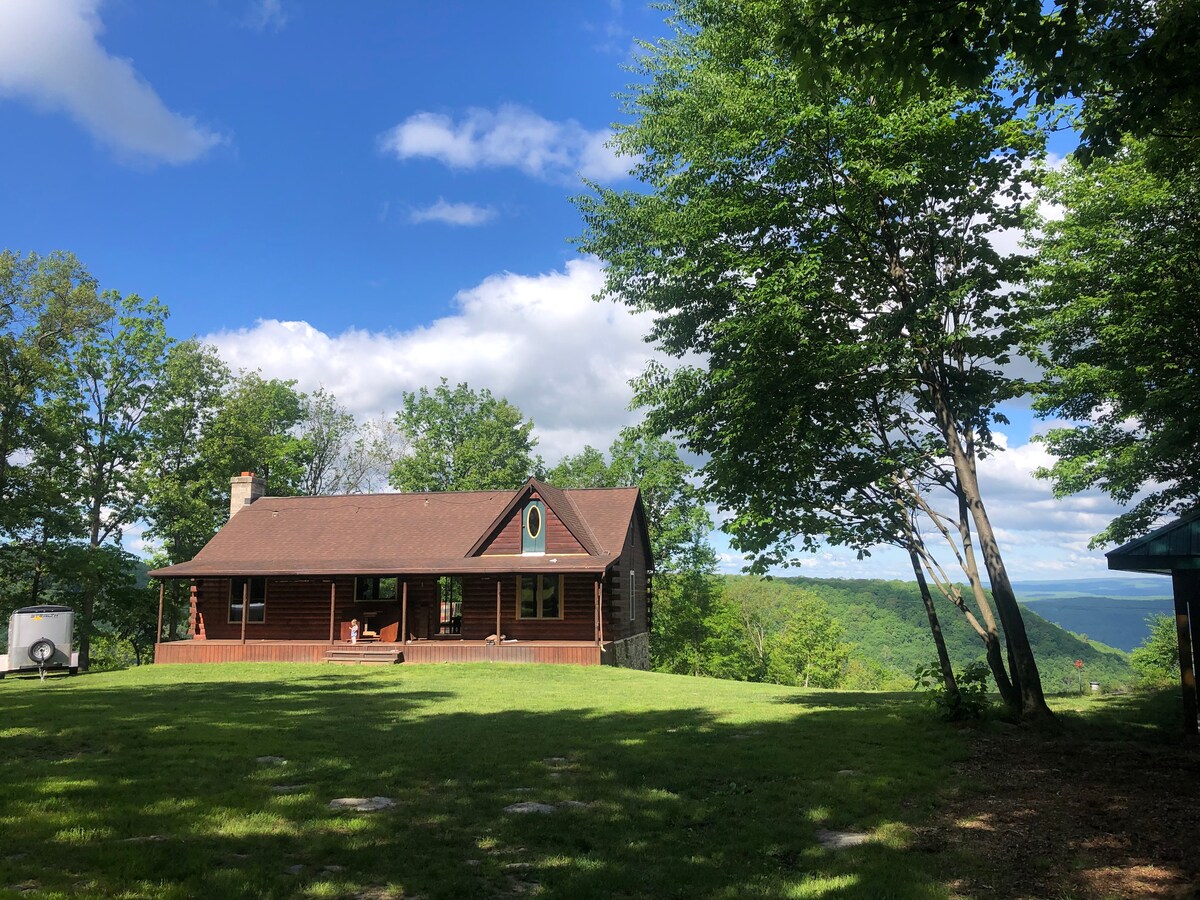 A spacious cabin is situated on a hillside, surrounded by green grass and trees. Large windows provide scenic views of the mountains in the distance, while a welcoming front porch invites relaxation in the outdoor space.