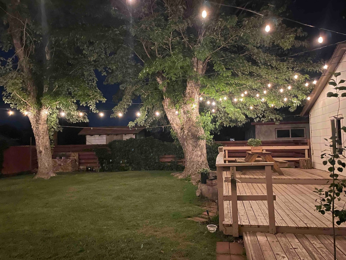 A spacious backyard is illuminated by warm string lights that hang between two large trees. A wooden deck extends into the yard, while a grassy area invites relaxation. The space is surrounded by greenery, providing a serene outdoor setting.