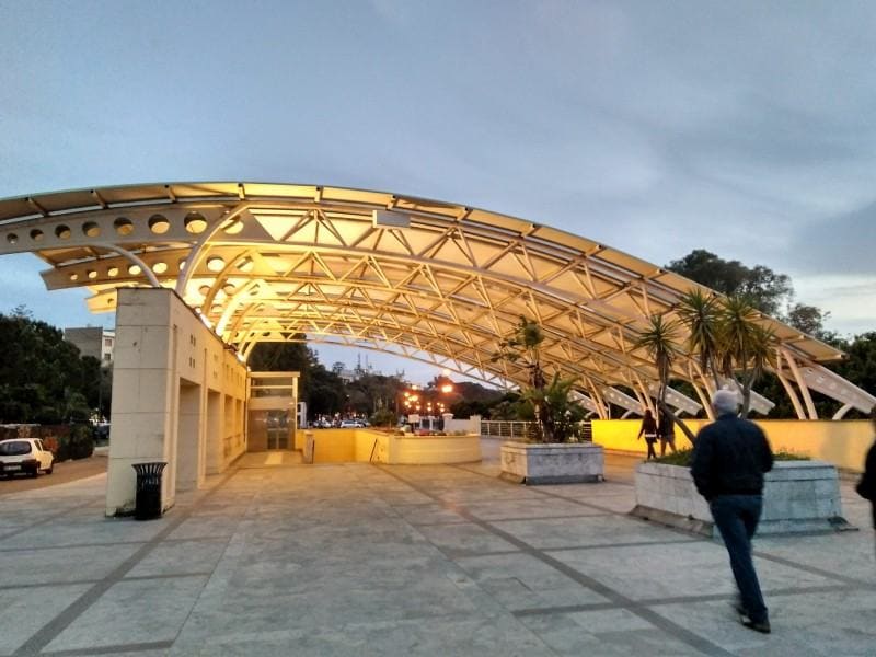 A modern architectural structure is featured, characterized by a large, curved canopy supported by metallic beams. Soft lighting illuminates the space, enhancing its design. Pathways are visible, with greenery and vehicles parked nearby, along with individuals walking through the area.