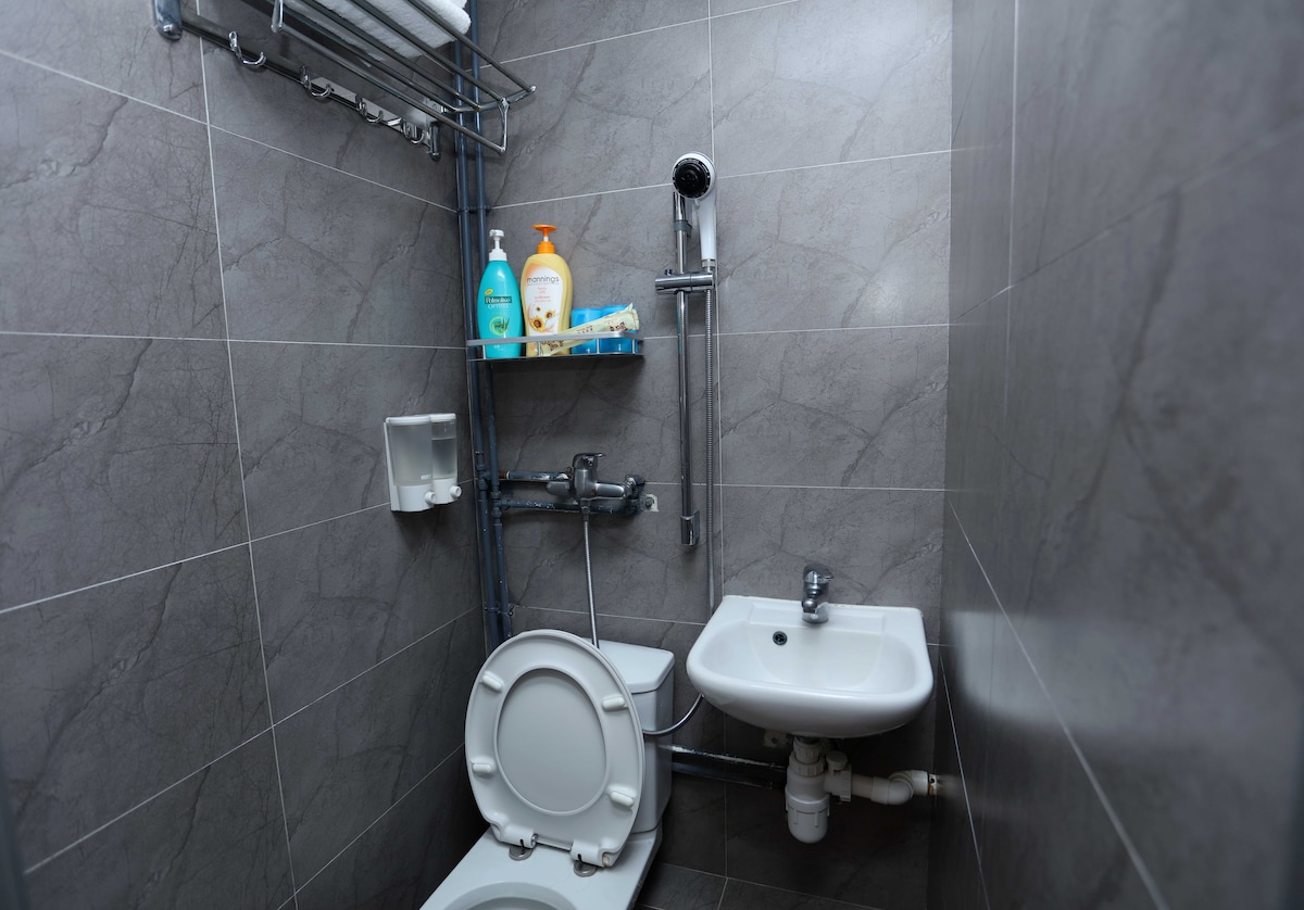 The compact bathroom is equipped with modern fixtures, including a toilet, a small sink, and a shower area. Soft lighting reflects off the grey tiled walls, enhancing the clean and functional design.