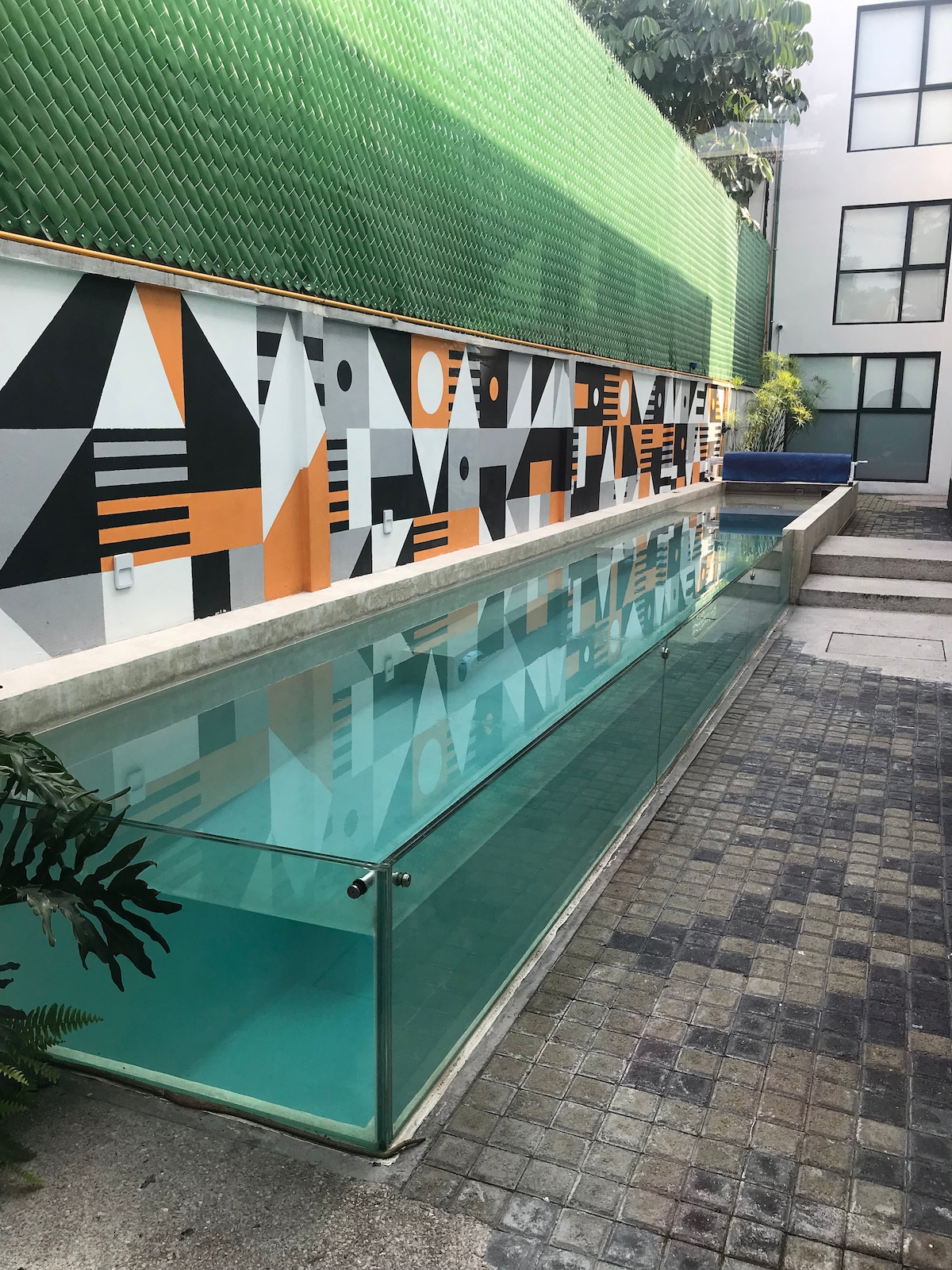 A modern swimming pool area is visible, featuring clear water surrounded by decorative tiles and artfully painted walls. The glass barrier offers a seamless view of the pool's surface. Lush greenery enhances the inviting atmosphere of the outdoor space.