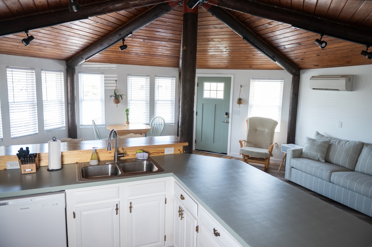 An open living area features a spacious kitchen with white cabinetry and modern appliances. A comfortable seating area includes a light-colored sofa and an armchair. Natural light fills the room through multiple windows, highlighting the wood beam ceiling and inviting atmosphere.