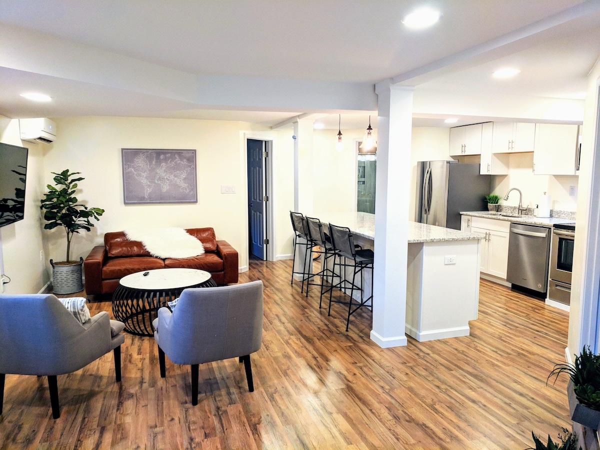 The open living space features a cozy seating area with two armchairs and a leather sofa. A modern kitchen with bar seating and stainless steel appliances is visible in the background, along with light hardwood flooring that connects the areas.