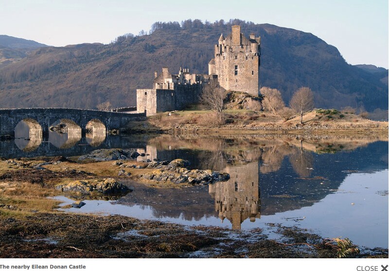 A historic castle is displayed on a rocky outcrop, reflecting in the calm waters of a loch. Surrounding hills are visible in the background, offering a serene natural setting. The stone bridge leading to the castle enhances the overall scenic landscape.