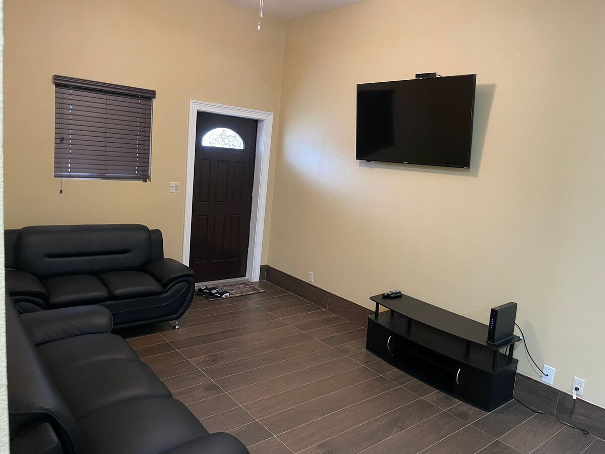A living area features a pair of sleek black sofas arranged around a low coffee table. A wall-mounted television is visible above a simple media console. Natural light enters through a window with blinds, complementing the earthy tones of the floor tiles.