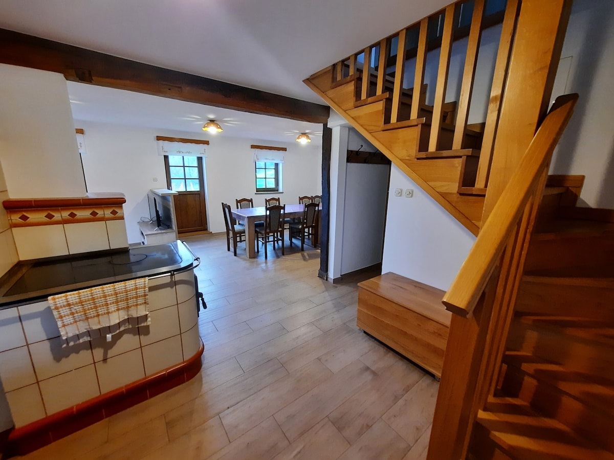 An open kitchen and dining area is featured, with a wooden dining table surrounded by chairs, illuminated by overhead lights. A staircase with wooden railings ascends to the upper level. Light wooden flooring and white walls contribute to a bright and airy atmosphere.