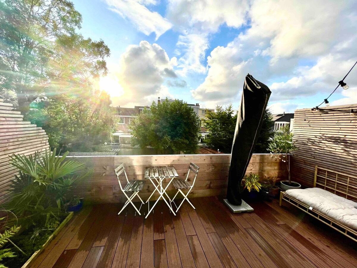 A spacious rooftop terrace features wooden decking surrounded by greenery. A small table with two chairs is placed for outdoor dining, while a lounge area with cushions is designed for relaxation. The scene is lit by warm evening sunlight, creating a tranquil ambiance.