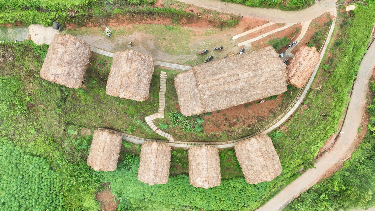 An aerial view displays a cluster of rustic structures featuring thatched roofs, arranged along a winding path. The surrounding landscape includes lush greenery and well-maintained pathways, highlighting a harmonious connection with nature.