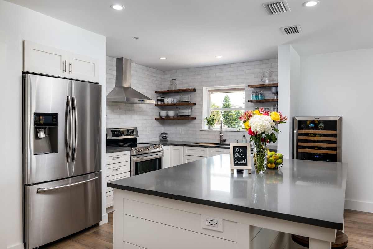 A modern kitchen is presented, featuring stainless steel appliances, including a refrigerator and oven. The countertop is gray, accompanied by a bouquet of flowers. Open shelves display stylish dishware, while a wine cooler is positioned on the side. Ample lighting enhances the clean, bright space.