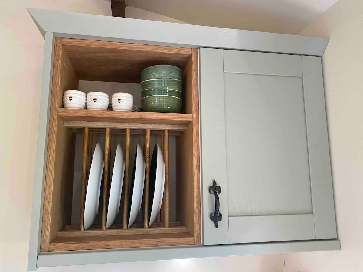 A wall-mounted cabinet displays neatly arranged dinnerware. White plates are positioned upright in a wooden rack, while smaller bowls and green plates are visible within the adjoining compartment. The cabinet features a pale green finish combined with natural wood accents.