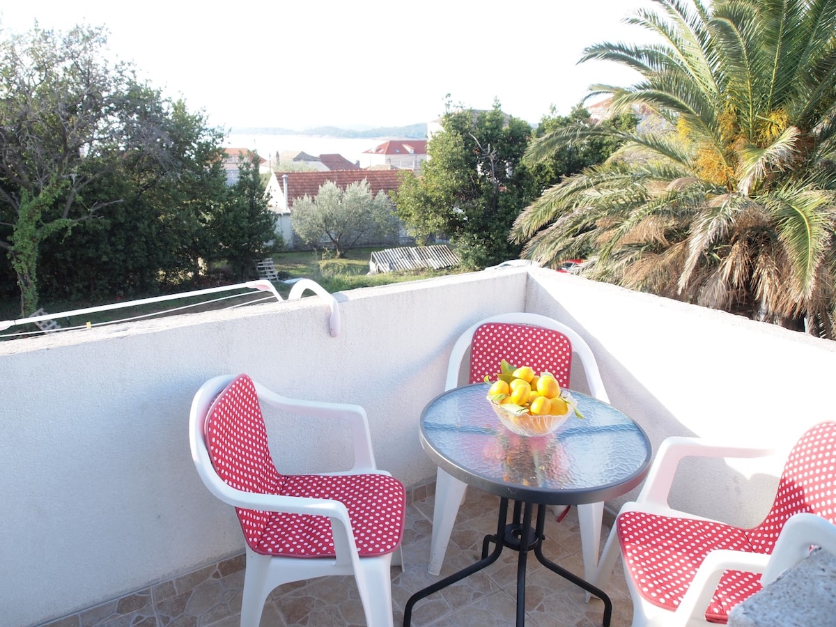 A small balcony features a round glass table surrounded by four white chairs with red polka dot cushions. A bowl filled with oranges and lemons sits at the center, while the sea and greenery can be seen in the background.