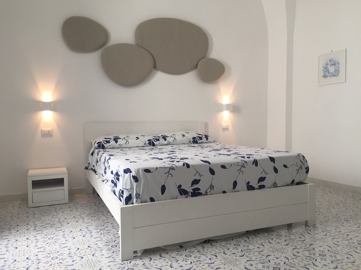 A spacious bedroom is furnished with a large bed dressed in floral-patterned linens. Soft lighting is provided by wall-mounted fixtures, and decorative wall art complements the neutral walls. Floor tiles feature a subtle pattern that enhances the room's serene ambiance.
